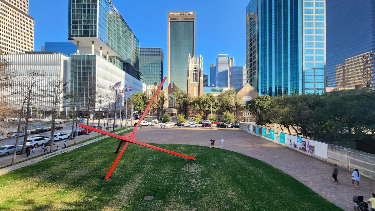 A green urban park with a red sculpture beneath downtown Dallas towers
