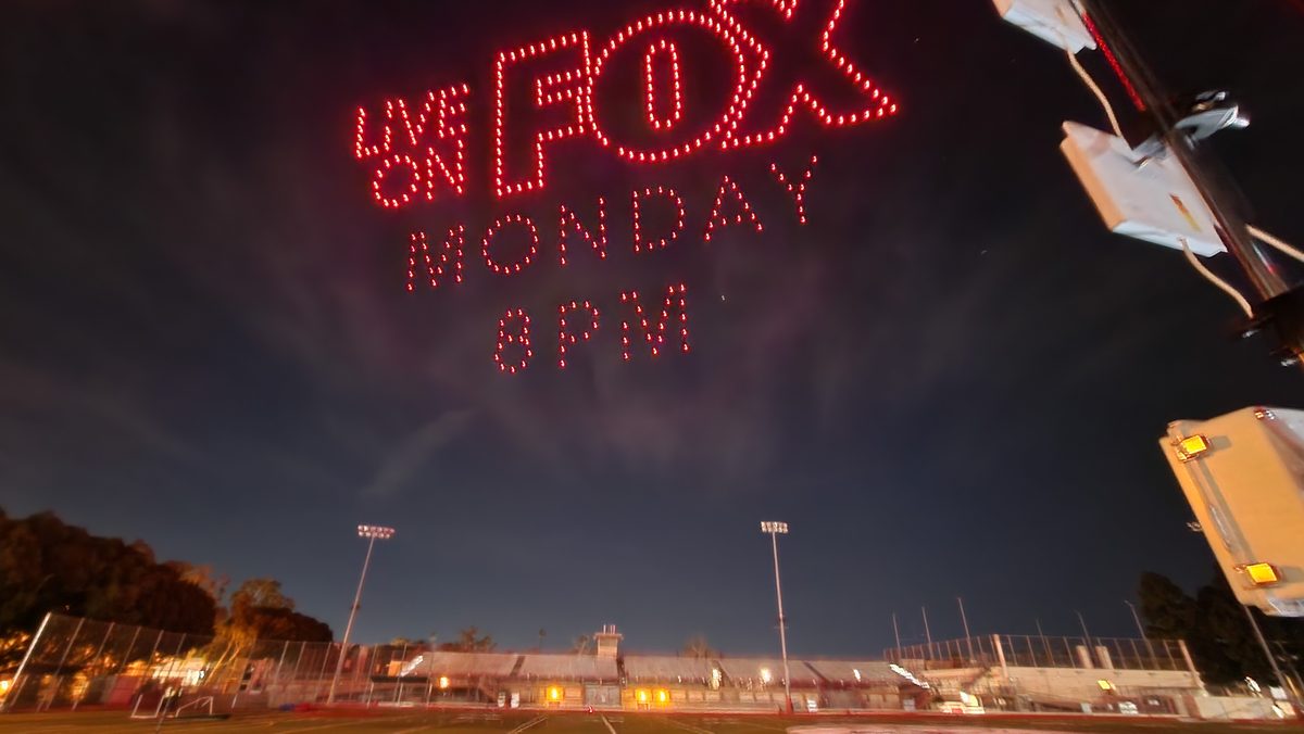 Red drones spelling a TV network promo in the night sky above a waterfront
