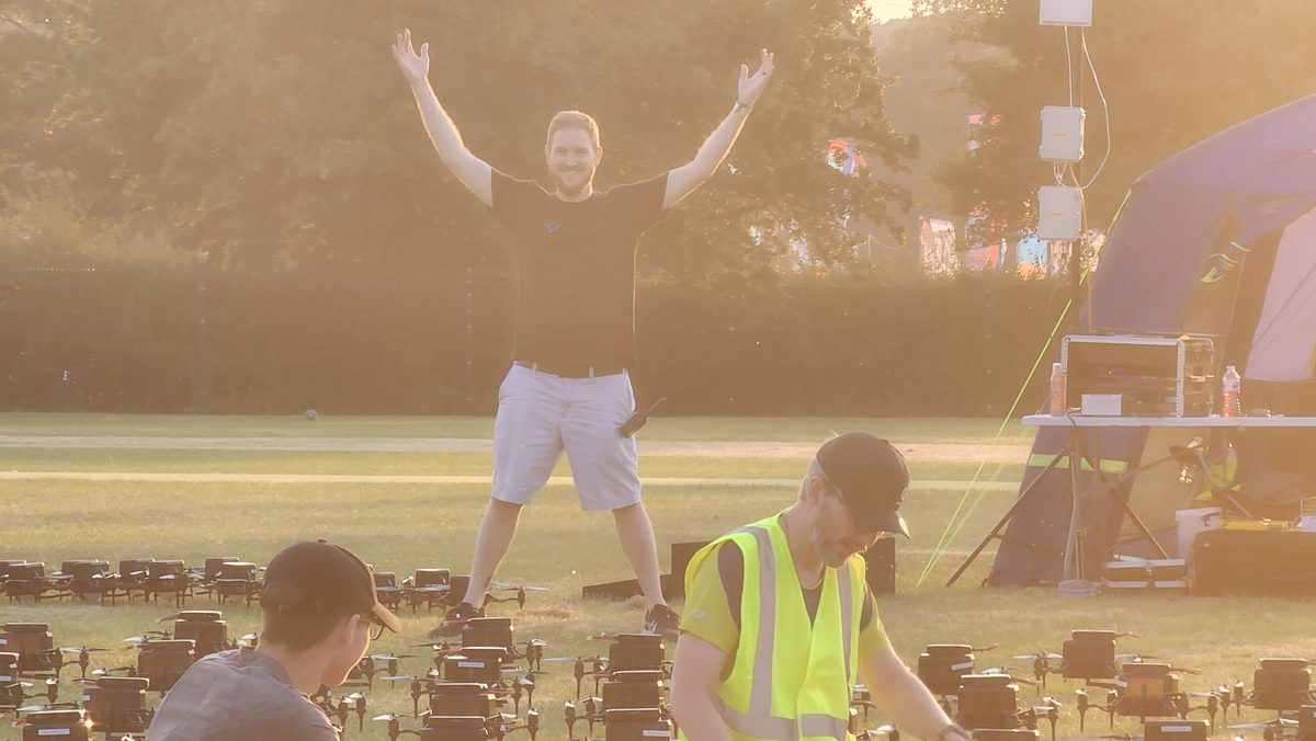 A drone show crew member celebrating with arms raised on a field of drones in golden haze