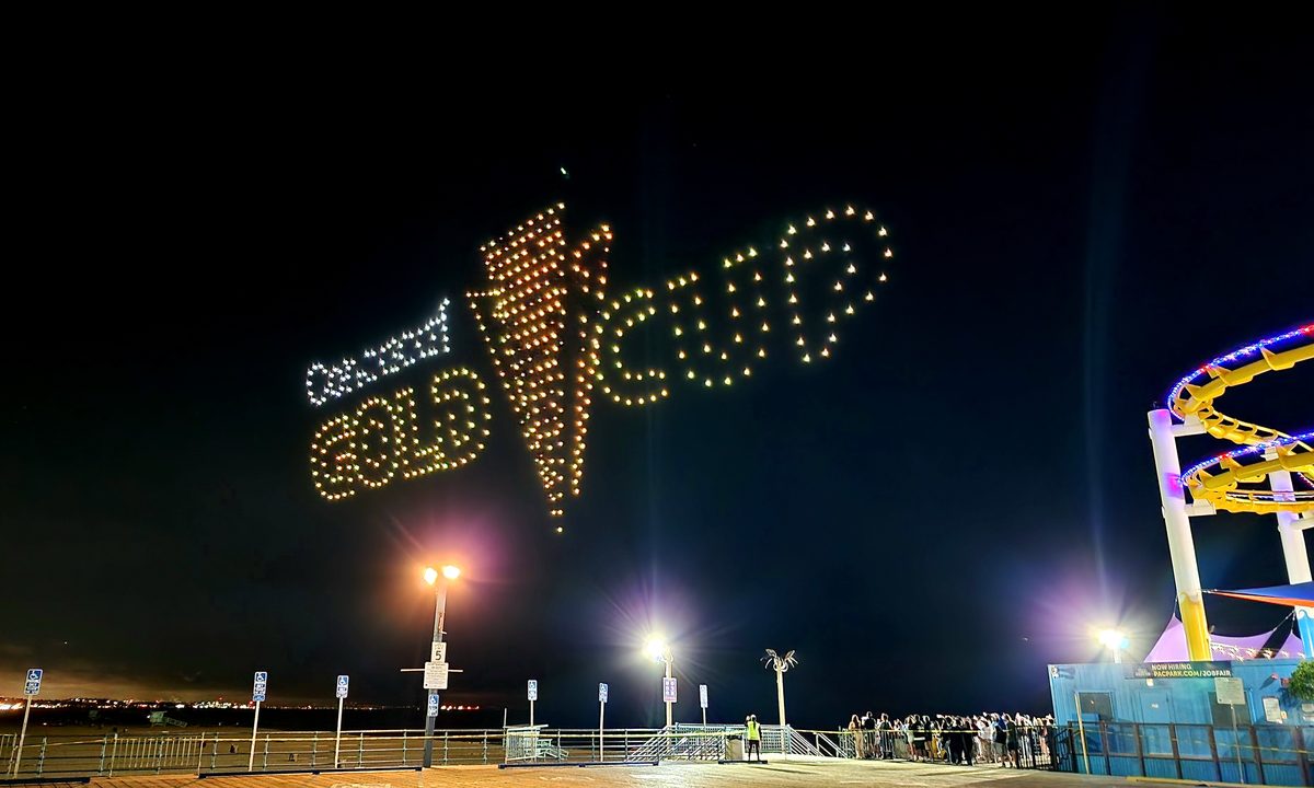 Drones forming a glowing trophy and text above a pier and Ferris wheel at night