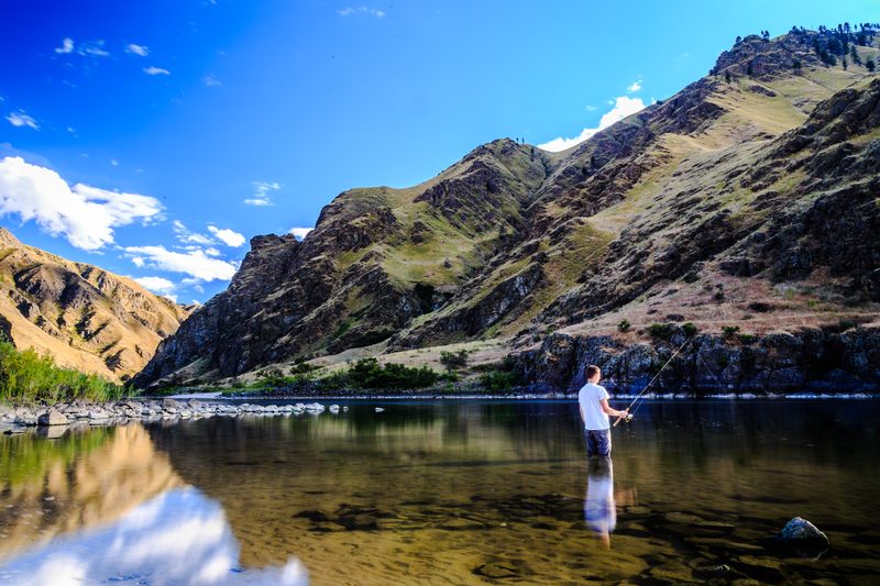 An angler standing in calm water below steep green canyon walls under a bright blue sky