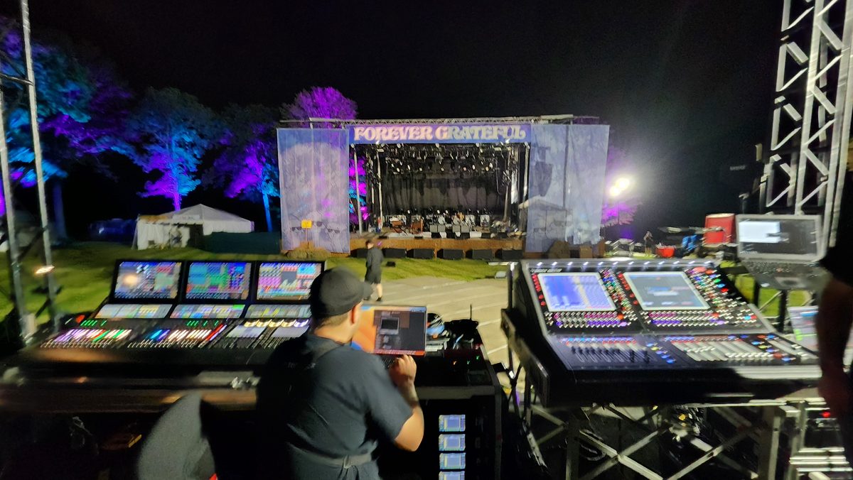 A sound engineer at a mixing desk facing a lit outdoor concert stage at night