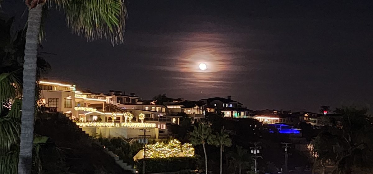 The moon rising over a harbor with lit homes along the shore
