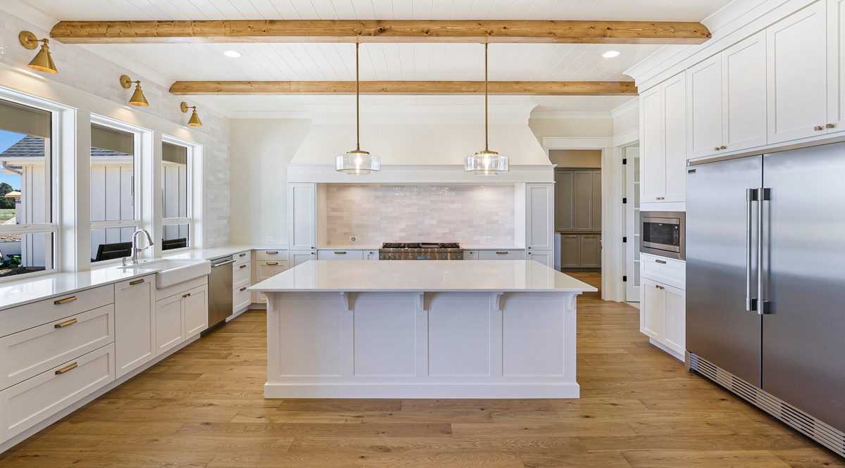 Long white kitchen with exposed wood ceiling beams, pendant lights and a stainless refrigerator