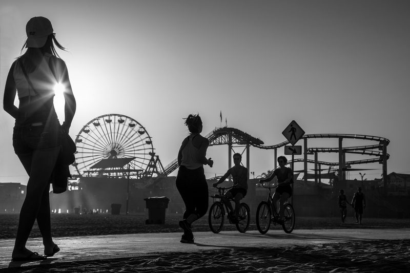 Black-and-white silhouettes of a runner and cyclists on a boardwalk in front of a pier Ferris wheel and roller coaster