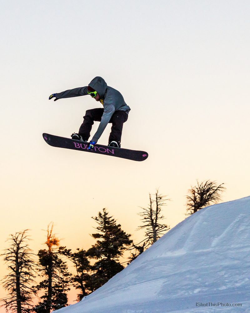 A snowboarder grabbing mid-air off a snow jump at sunset with pine trees below