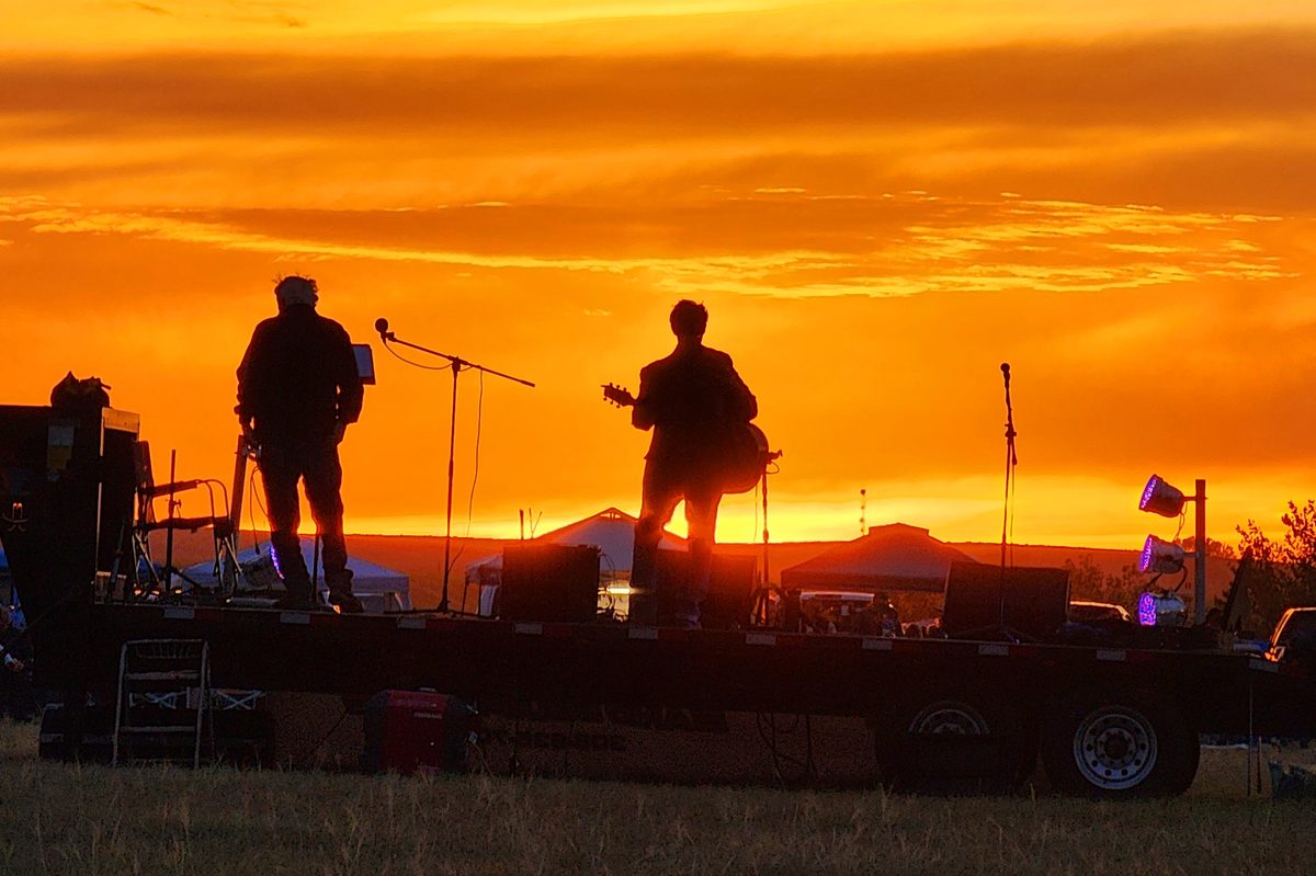 Musicians silhouetted on a small outdoor stage against an orange sunset