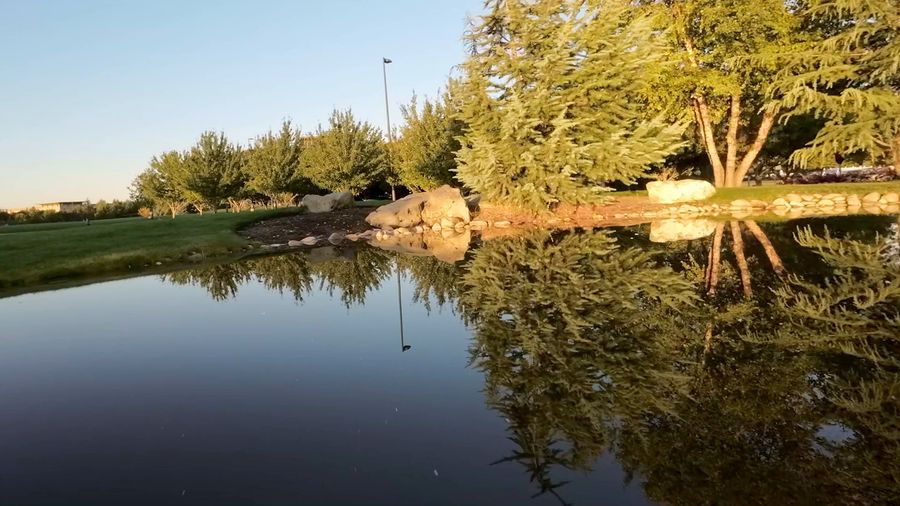 FPV drone weaves low through office-campus gardens, past a pond, flower beds and stone walls in golden light