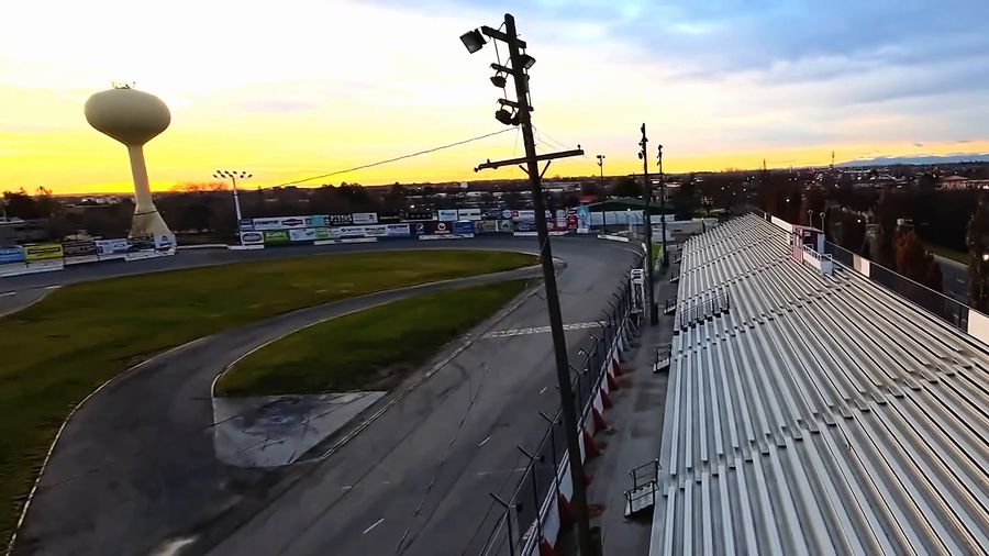 Drone swoops around an empty oval race track and grandstand at sunset, the Meridian water tower behind it