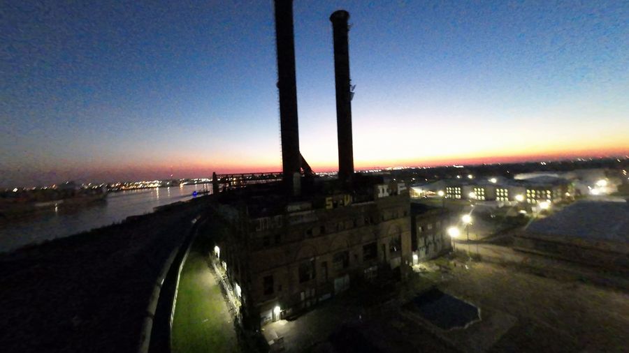 FPV flight around the two tall chimneys of an old riverside power plant at dusk, city lights beyond