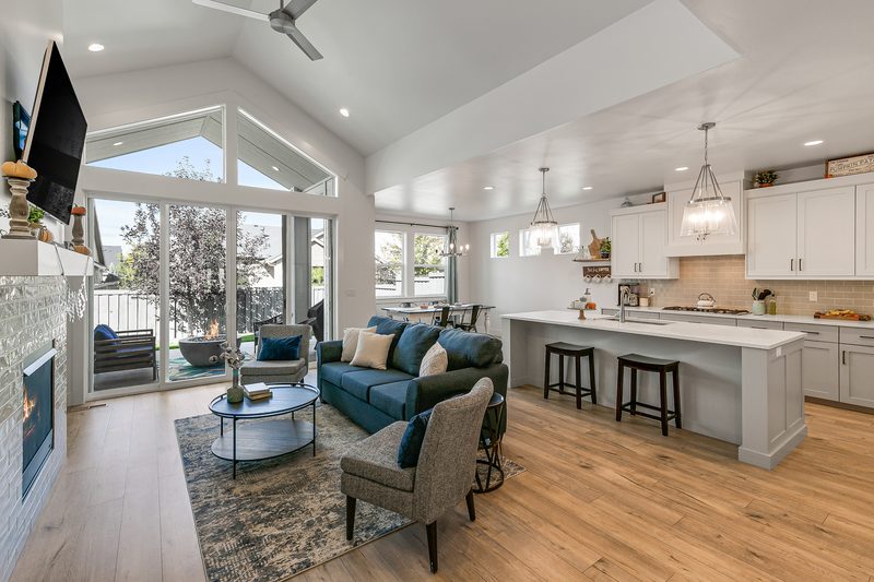 Delivered listing photo: bright vaulted living room open to a white kitchen with an island, with the backyard visible through the tall windows